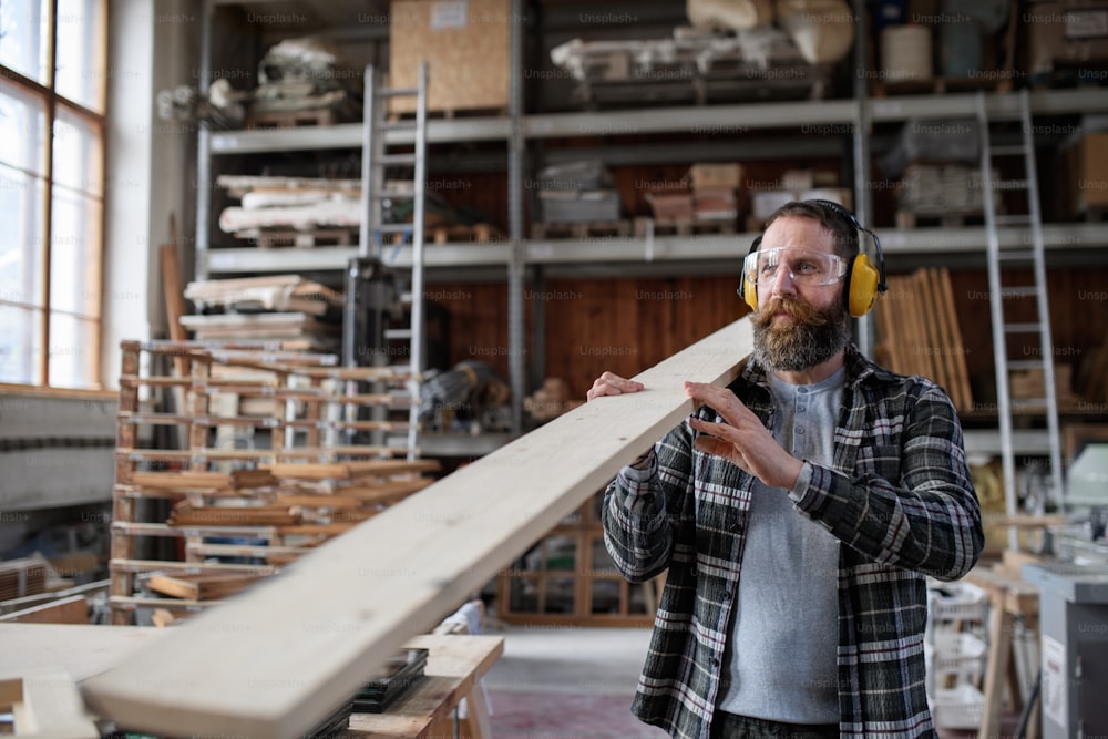 Ein reifer männlicher Tischler mit Gehörschutz trägt Holzbrett, drinnen in der Schreinerei. Konzept für kleine Unternehmen.