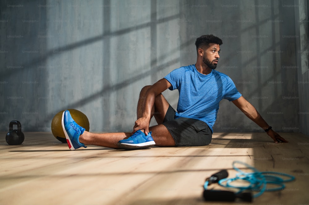 A young African American sportsman sitting on floor and doing stretching exercise indoors, workout training concept.