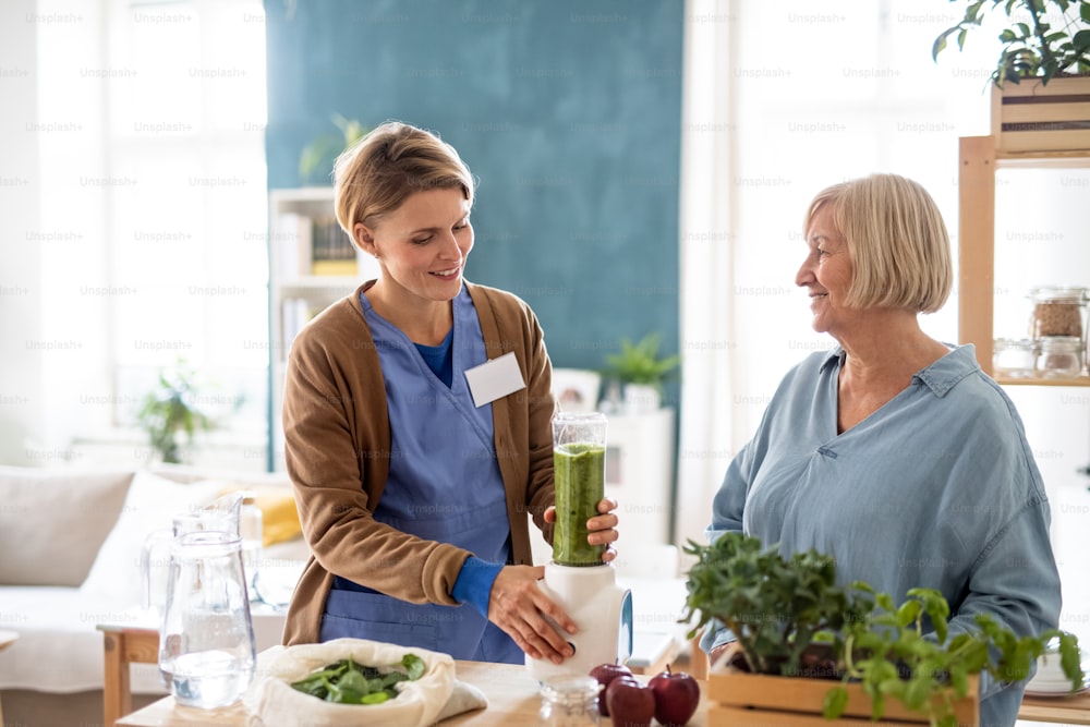 Senior woman with caregiver or healthcare worker indoors, preparing healthy smoothie drink.