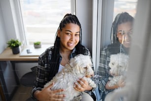 Ein Porträt eines glücklichen Teenagermädchens mit Hund, das zu Hause drinnen sitzt und in die Kamera schaut.