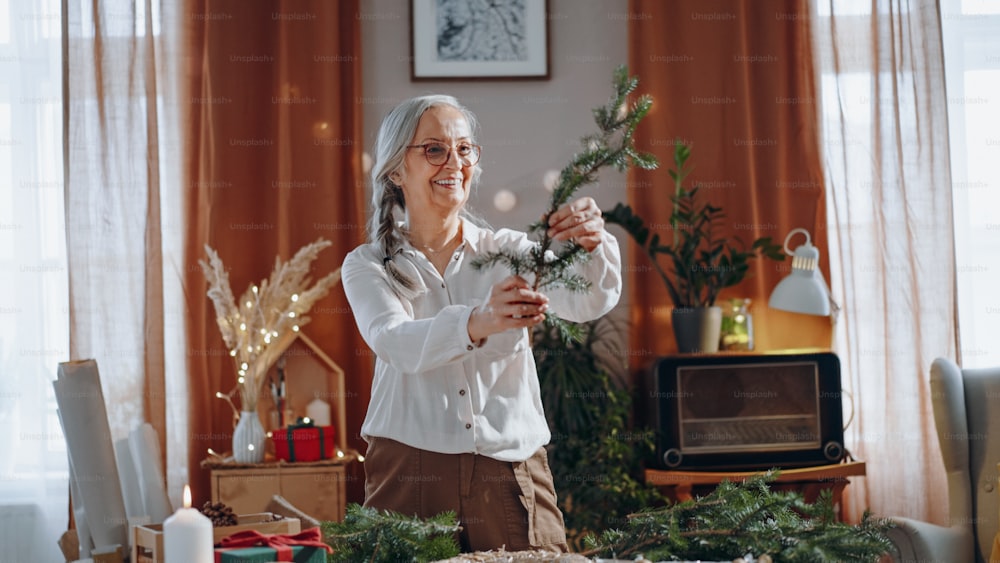 Una mujer mayor feliz cortando una rama para hacer una corona de Navidad en el interior de casa