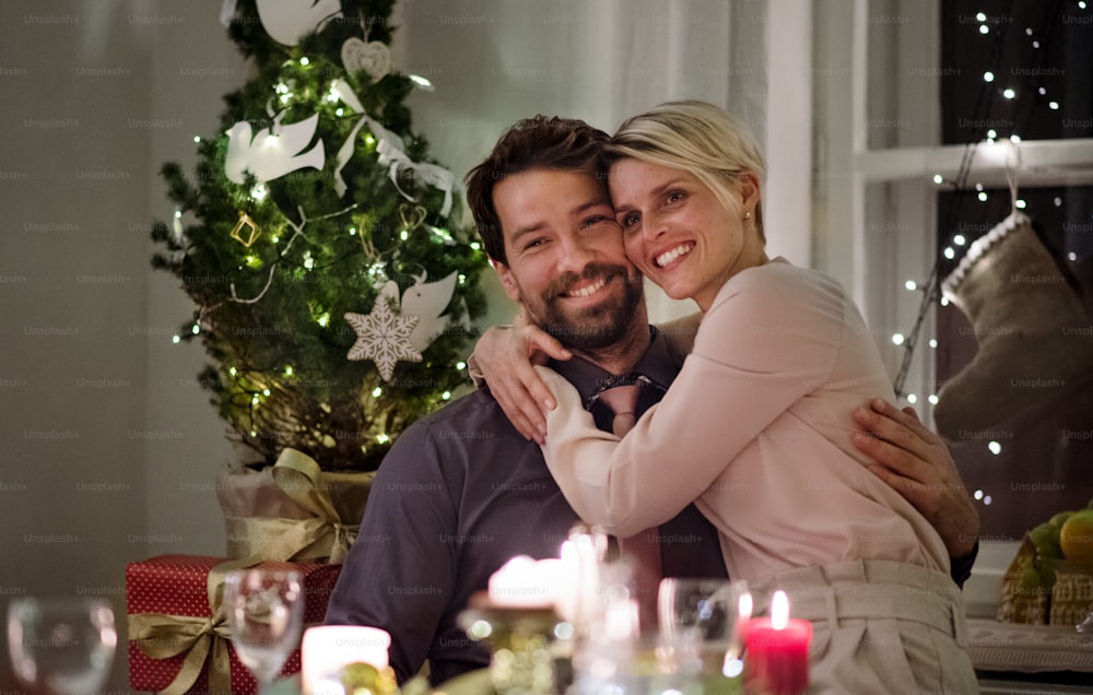 Vista frontal de una pareja feliz sentada en el interior de la mesa celebrando la Navidad, abrazándose.