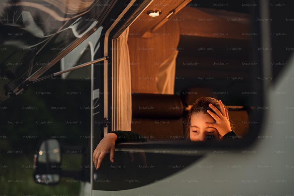 Uma menina pequena feliz olhando para fora através da janela da caravana ao anoitecer, viagem de férias em família.