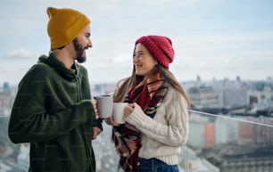 A young couple with coffee standing and looking at each other outdoors on balcony with urban view.