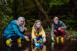 A small girl with mother and grandmother playing with paper boats in lake outoors in nature.