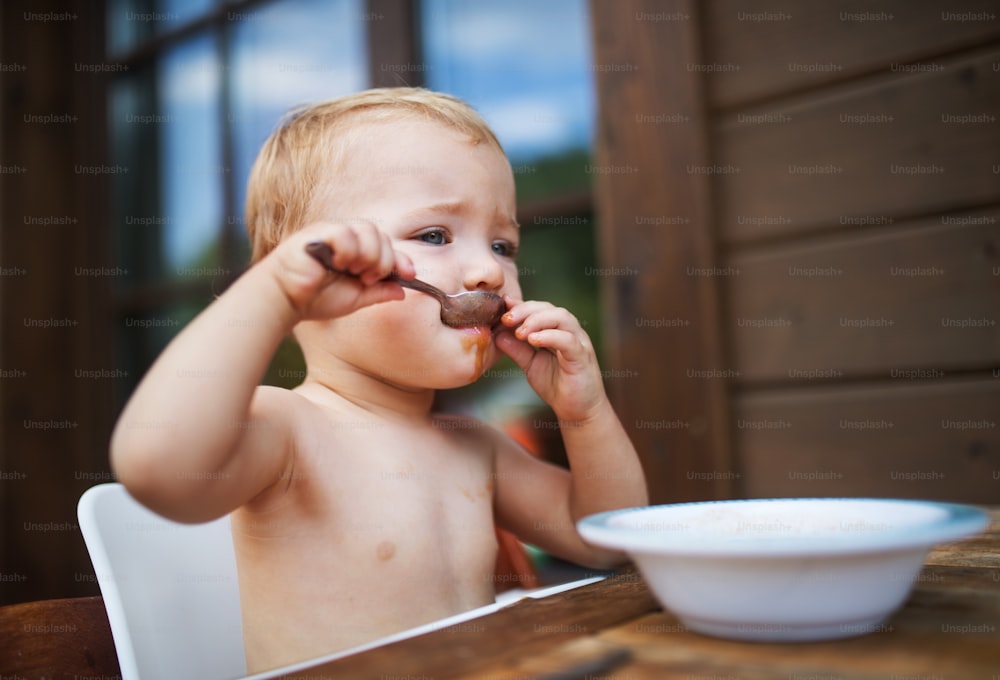 Cute topless small girl standing by a table on a patio in summer, eating soup.