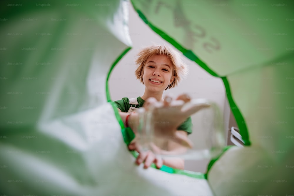 An image from inside green recycling bag of girl throwing a glass bottle to recycle.
