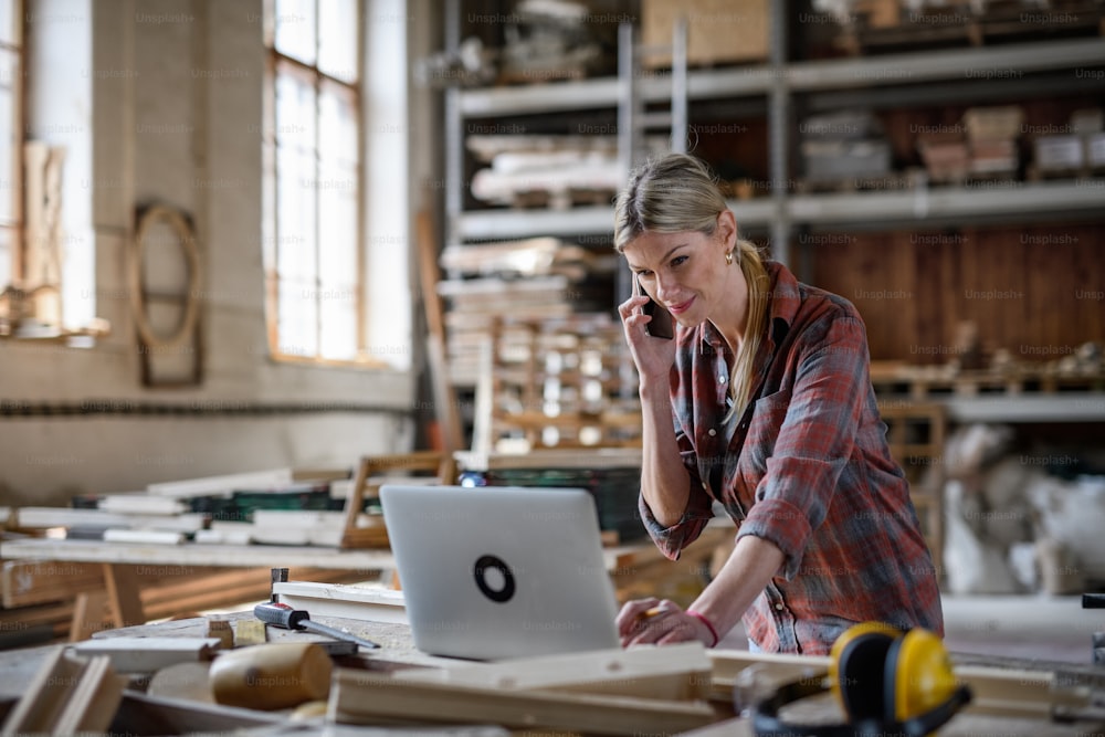 Uma mulher adulta média usando laptop e smartphone dentro de casa na oficina de carpintaria, conceito de pequena empresa.