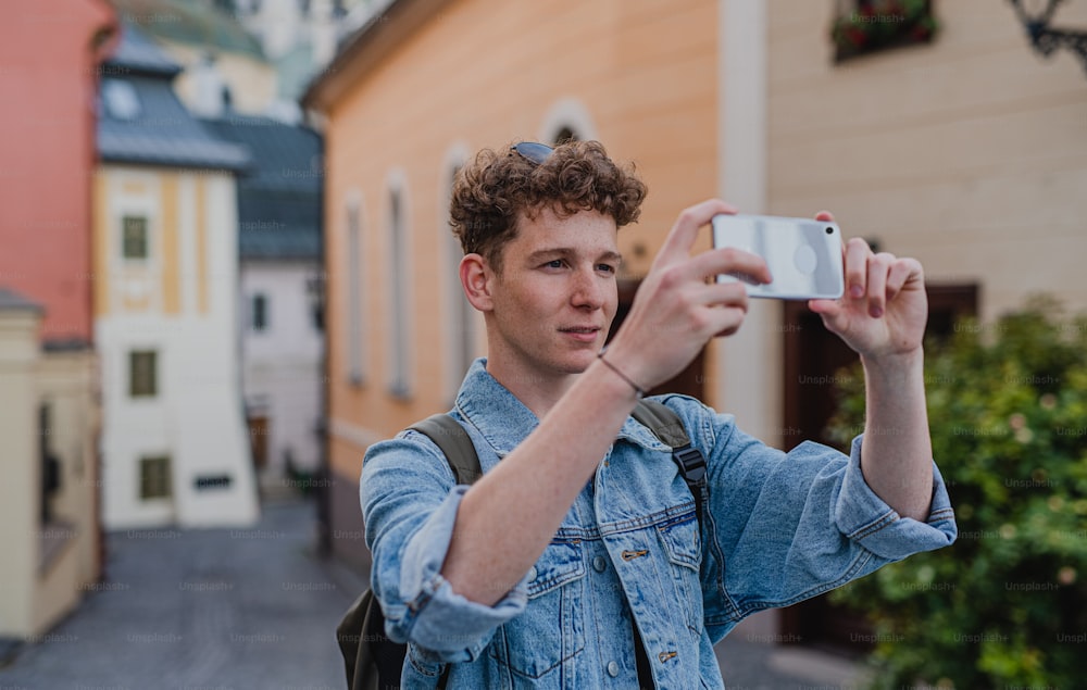 A young man outdoors on trip in town, taking photograph with smartphone.