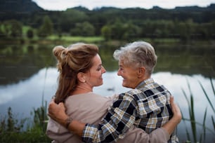A rear view of happy senior mother embracing with adult daughter when sitting by lake outdoors in nature