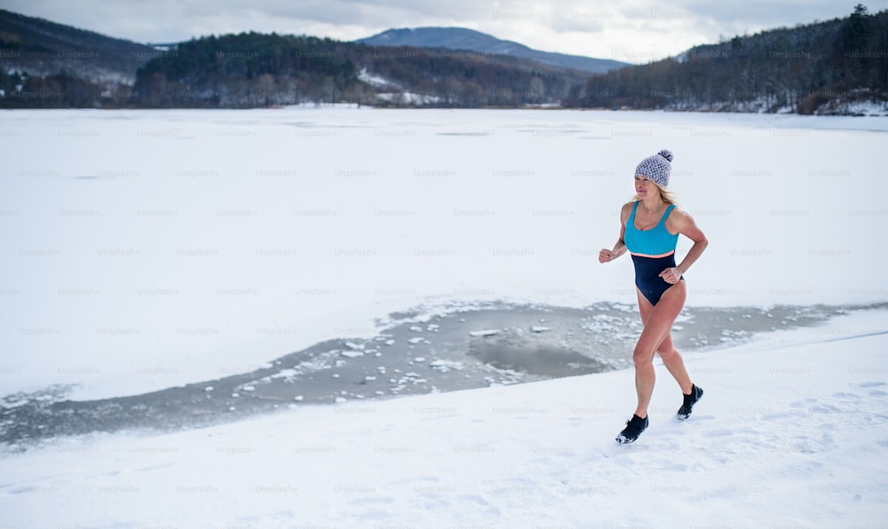 Happy active senior woman in swimsuit running outdoors in winter, cold therapy concept.