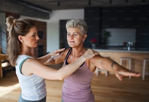 Una mujer mayor activa en ropa deportiva haciendo ejercicio con una fisioterapera en el interior de su casa.