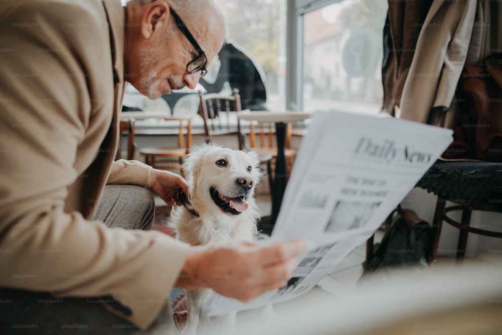 A happy senior man sitting in cafeteria and reading newspaper with his dog.