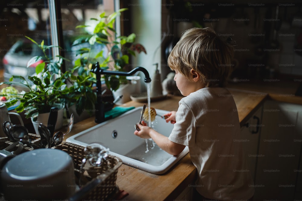 A small boy using eco friendly brush to wash dishes indoors at home, daily chores concept.