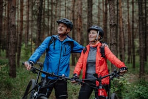 A senior couple bikers with e-bikes admiring nature outdoors in forest in autumn day.