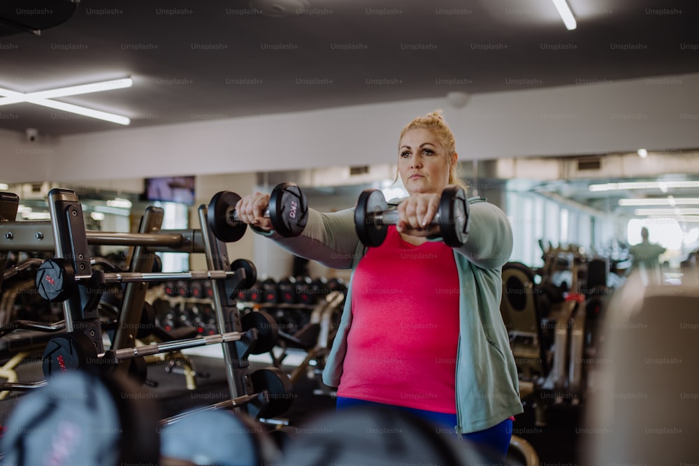 A plus size woman training and lifting dumbbells indoors in gym