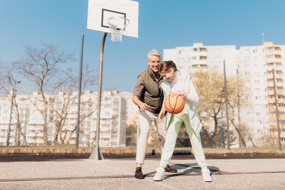 A happy father and teen daughter playing basketball outside at court.