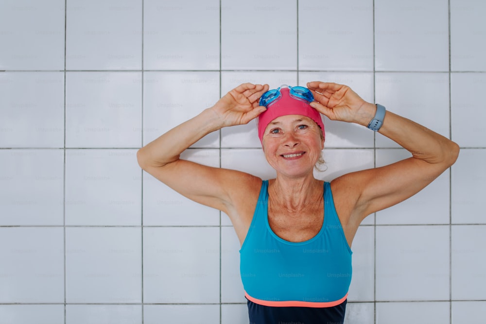 An active senior swimmer woman standing against white tiles wall in swimming pool, looking at camera.