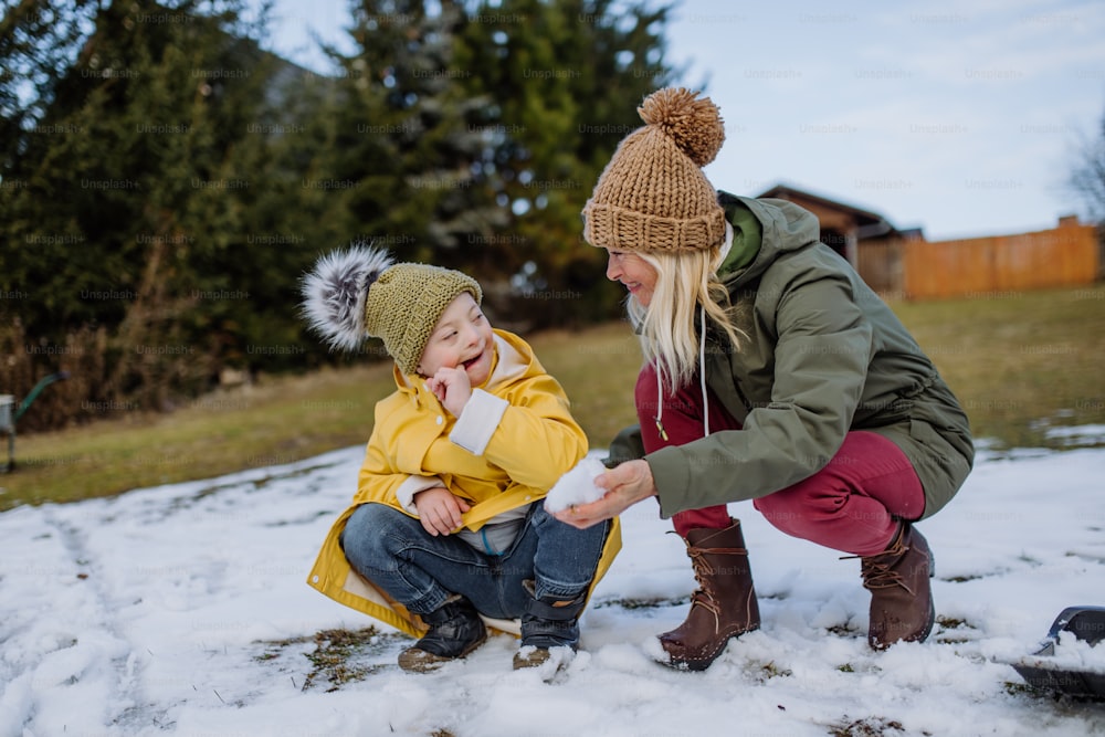 A boy with Down syndrome with his mother playing with snow in garden.