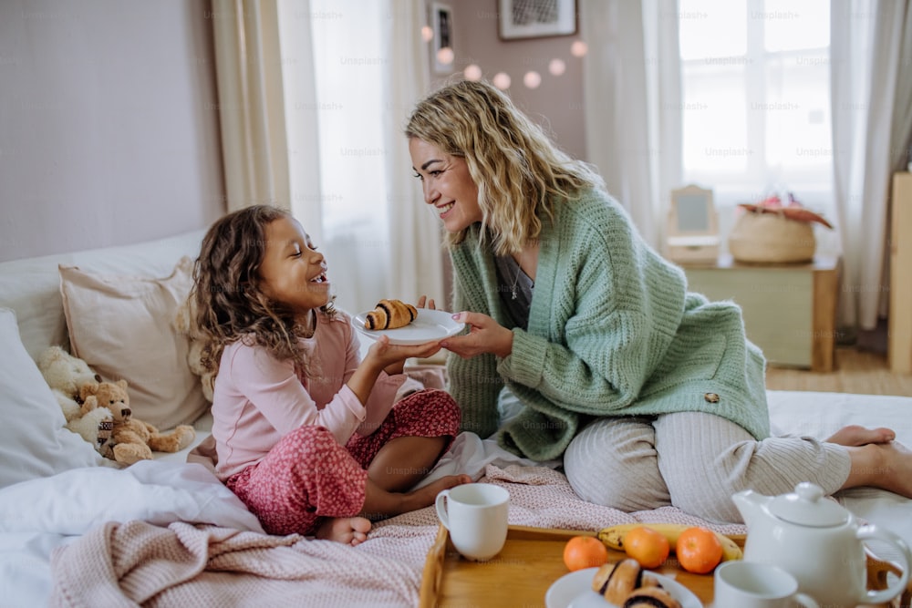 A happy mother with her little daughter having breakfast together in bed at home.