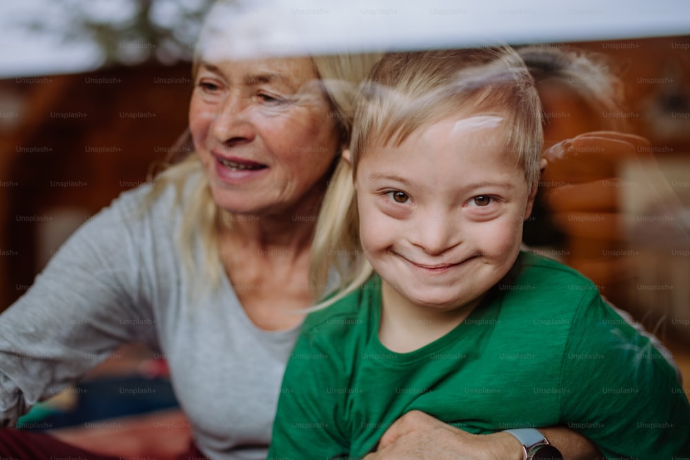 Um menino com síndrome de Down com sua avó olhando para a câmera através da janela em casa.