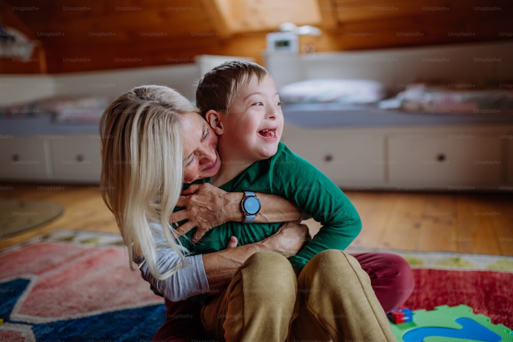 Ein glücklicher Junge mit Down-Syndrom, der auf dem Boden sitzt und sich mit seiner Großmutter zu Hause umarmt