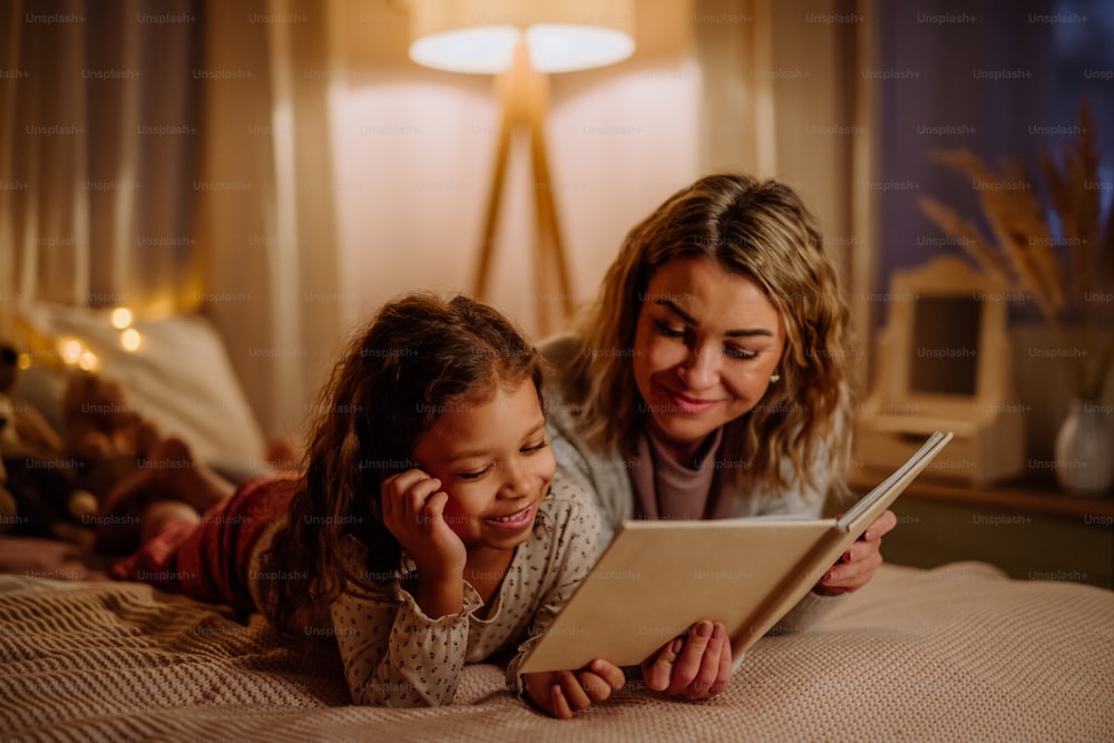 A happy mother with her little daughter lying on bed and reading book in evening at home.