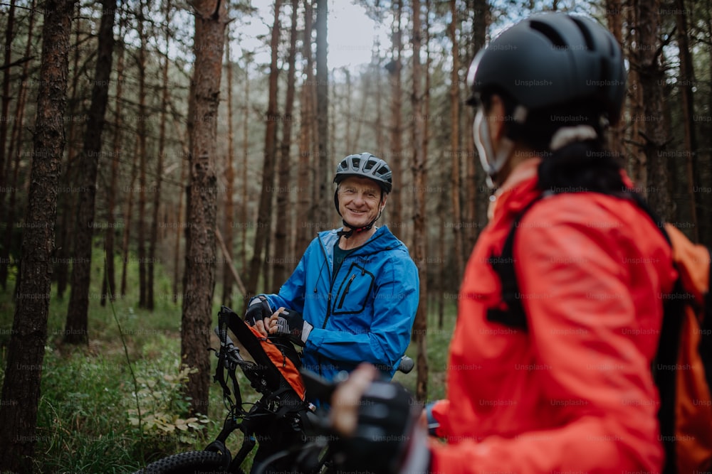 An active senior couple having break and talking when riding bikes outdoors in forest in autumn day.