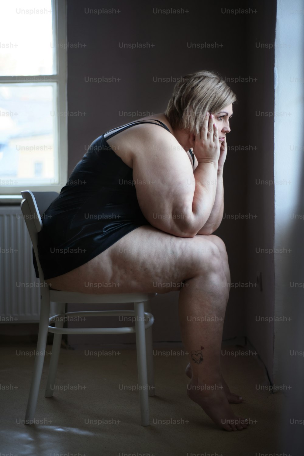 Mulher gorda solitária deprimida sentada e olhando pela janela em casa no escuro.