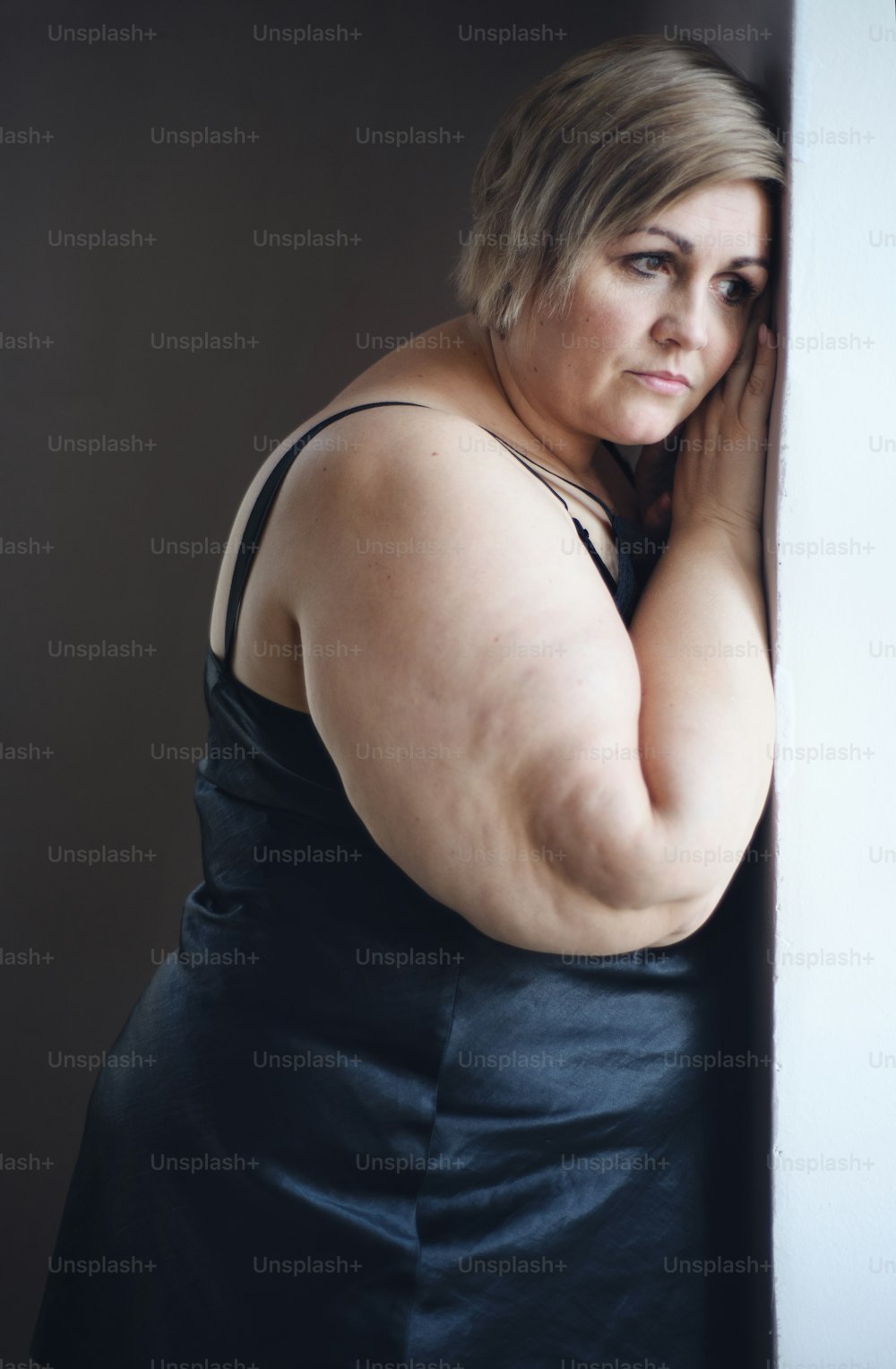 우울한 외로운 뚱뚱한 여자가 서서 집에서 창문을 통해 바라보고 있습니다.