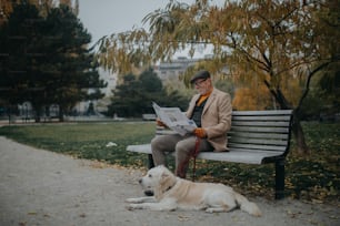 A happy senior man sitting on bench and reading newspaper during dog walk outdoors in park in city.