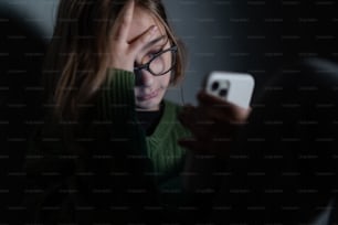 Uma garotinha triste, sozinha na escuridão, sentada e usando smartphone.