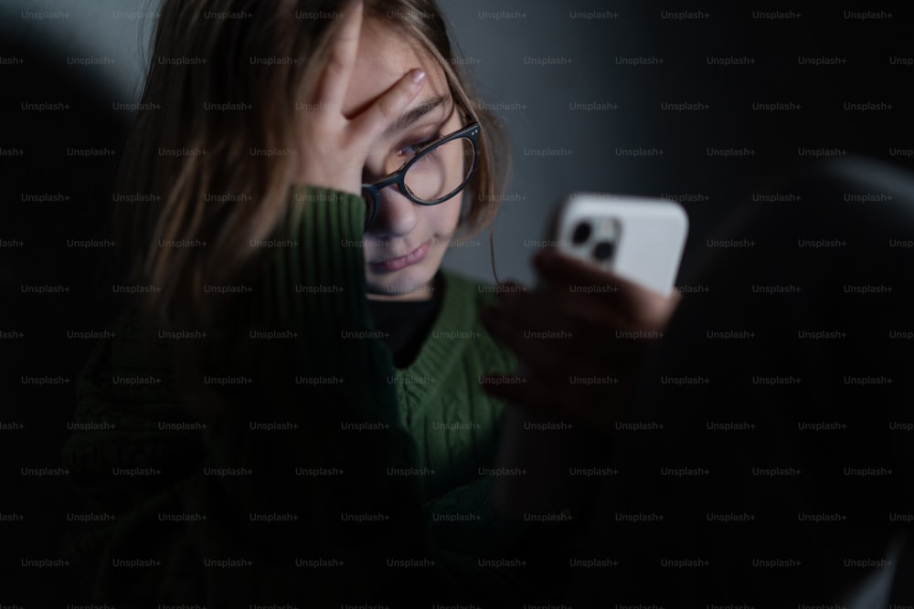 Una niña triste, sola en la oscuridad, sentada y usando un teléfono inteligente.