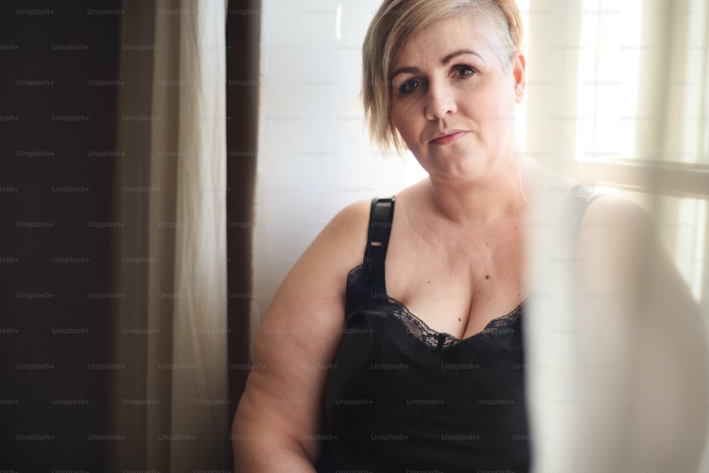 An overweight woman in underwear sitting by window and looking at camera.