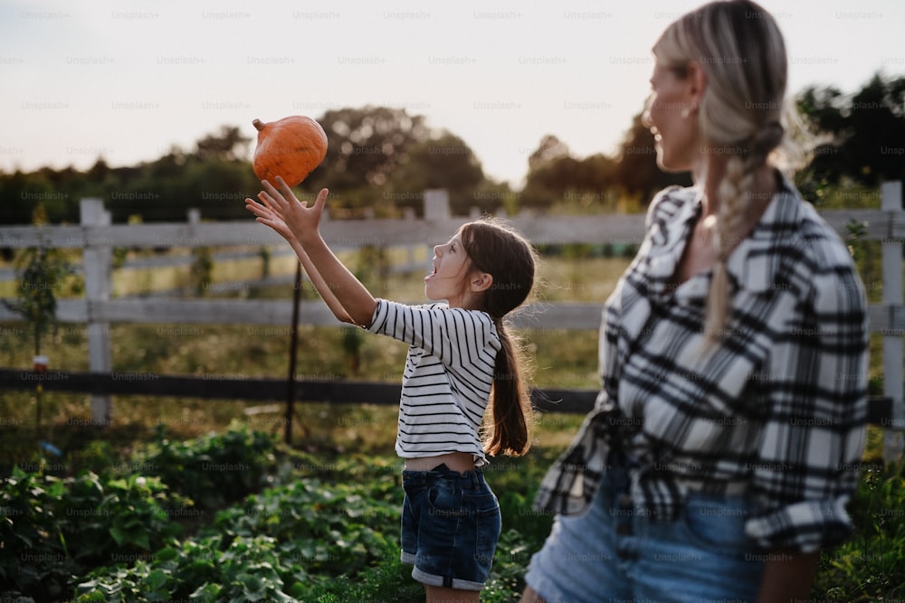 A happy little farmer girl throwing pumpkin with mother outdoors at garden.
