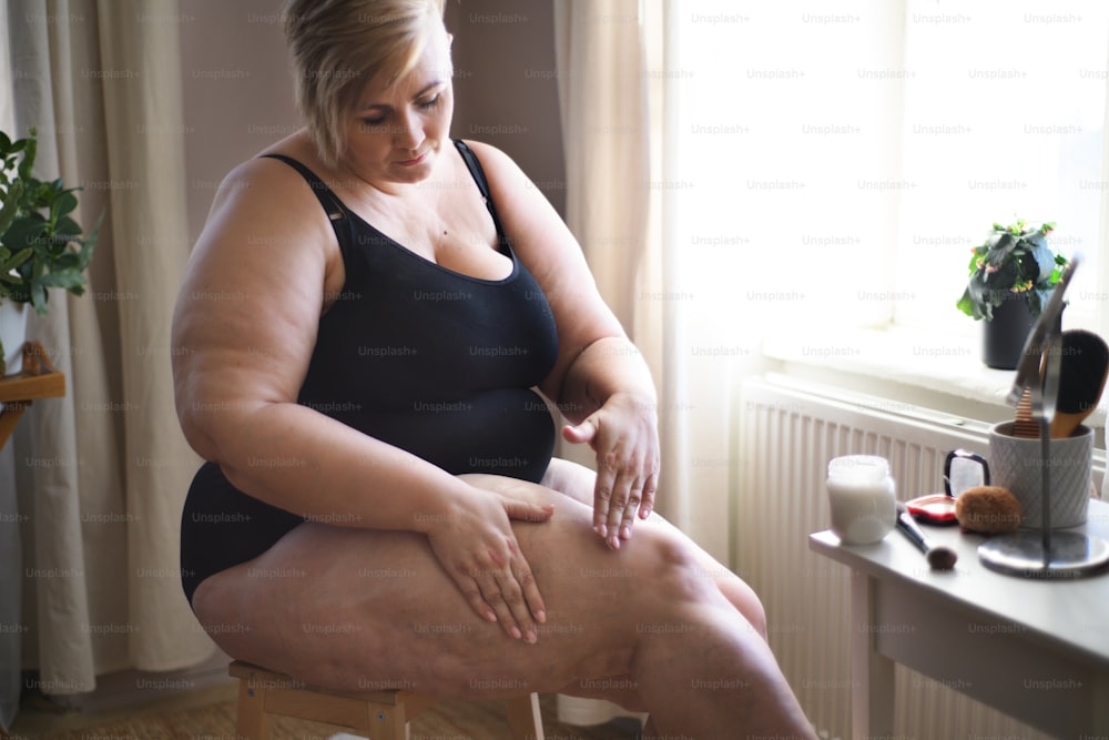 Una mujer gorda sentada y aplicándose crema en las piernas en casa, concepto de cuidado personal.