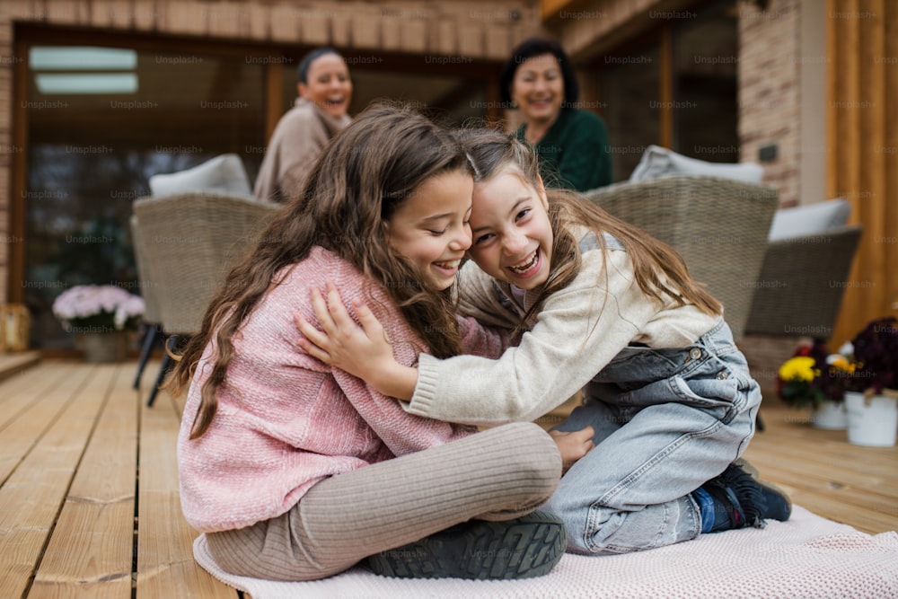 Two happy little sisters hugging outdoors in a patio in autumn, mother and grandmother at background.