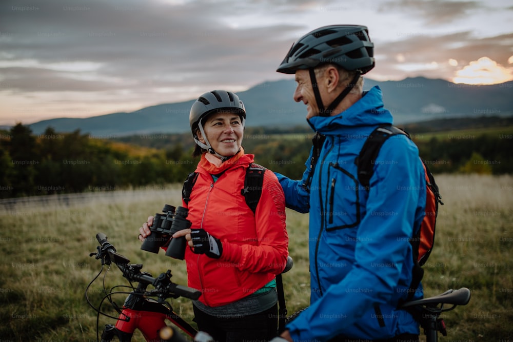 A senior couple bikers with binoculars admiring nature outdoors in meadow in autumn day.