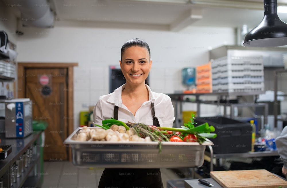 A female chef carrying tray with fresh vegetables and looking at camera in restaurant kitchen.