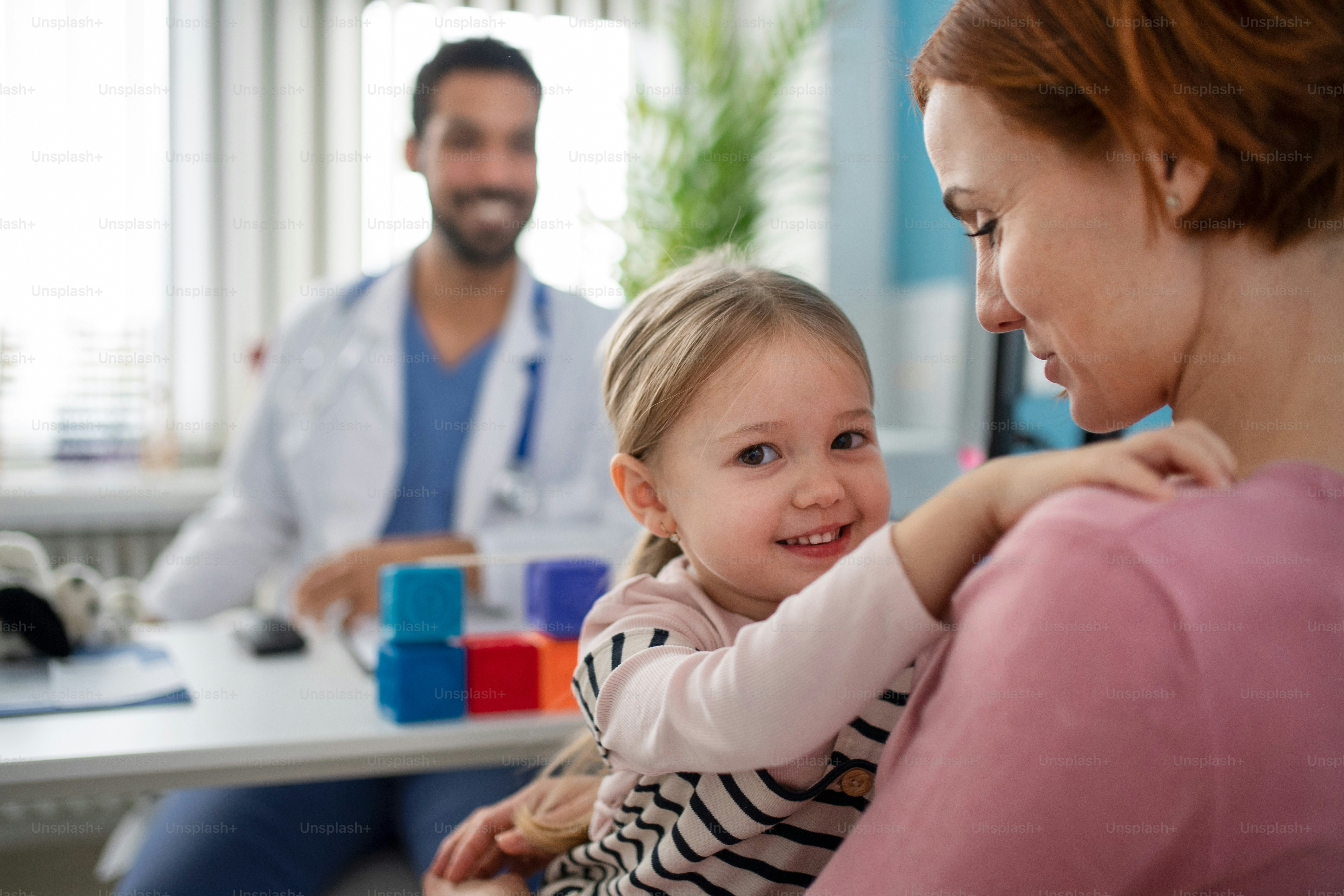 Ein kleines Mädchen mit ihrer Mutter in der Arztpraxis auf Konsultation, in die Kamera schauend.