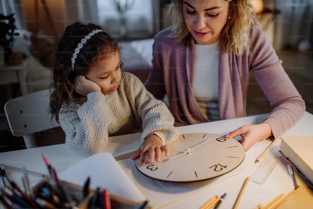 A happy little girl learning time with wooden clock with her mother in evening at home.