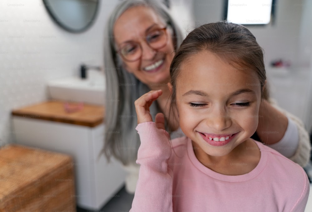 A happy senior grandmother and granddaughter standing indoors in bathroom, brushing hair in morning.