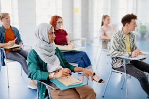 Ein Porträt einer multiethnischen Gruppe von Universitätsstudenten, die drinnen im Klassenzimmer sitzen und studieren.