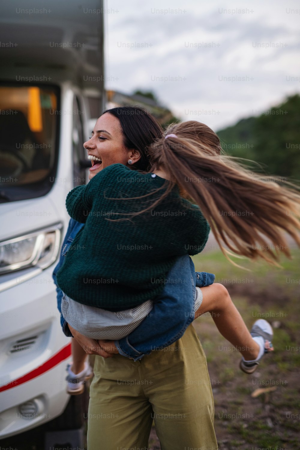 Happy mother with daughter having fun by car outdoors in campsite at dusk, caravan family holiday trip.