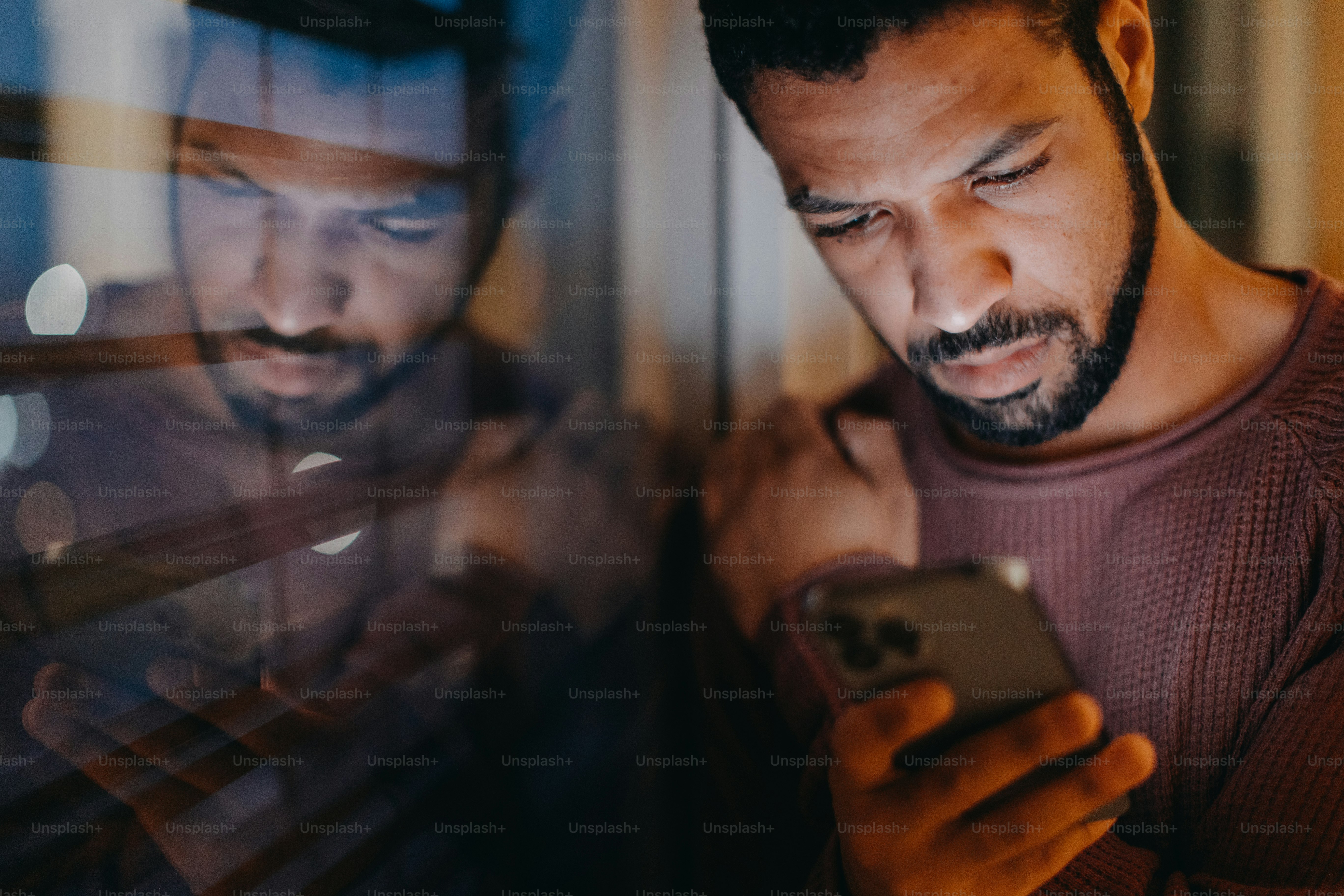 A young man with a smart phone looks sad, standing by window in evening at home