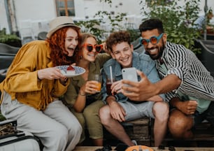 街歩きの屋外カフェに座って自撮りをしているスマートフォンを持った幸せな若者のグループ。