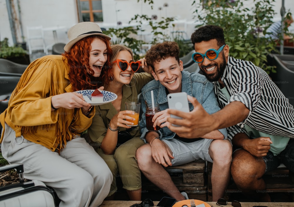 街歩きの屋外カフェに座って自撮りをしているスマートフォンを持った幸せな若者のグループ。