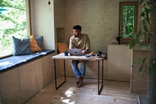 Un joven feliz con una computadora portátil y un teléfono inteligente sentado, descansando en el interior en una casa en el árbol, un fin de semana y un concepto de oficina remota.