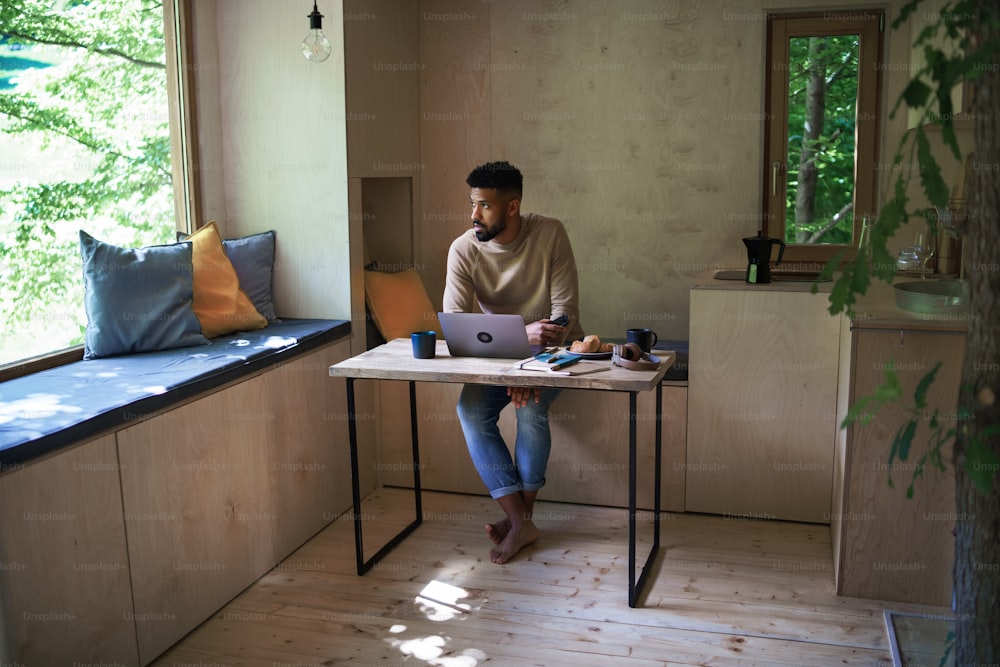 Ein glücklicher junger Mann mit Laptop und Smartphone sitzt, ruht sich drinnen in einem Baumhaus, Wochenendausflug und Remote-Office-Konzept aus.