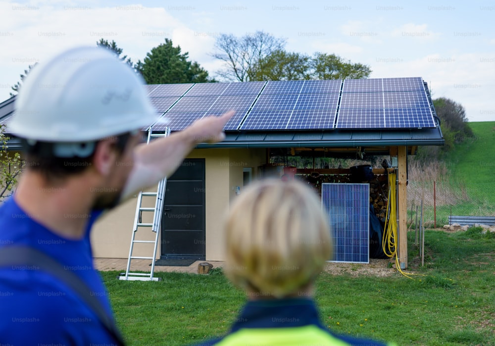 Un uomo e una donna installatori solari ingegneri con tablet durante l'installazione del sistema di pannelli solari sulla casa, vista posteriore.